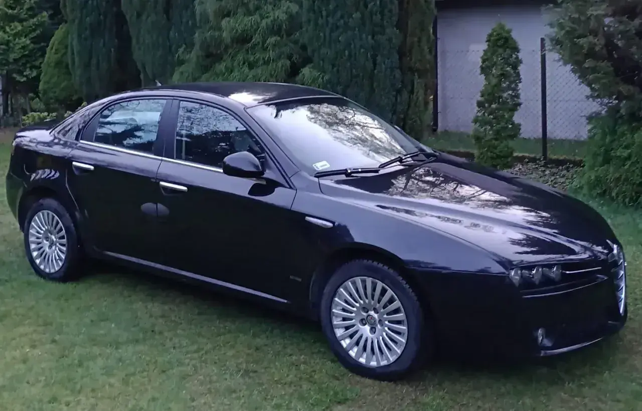 Czarna Alfa Romeo 159 lśni na trawie, otoczona zielenią drzew.