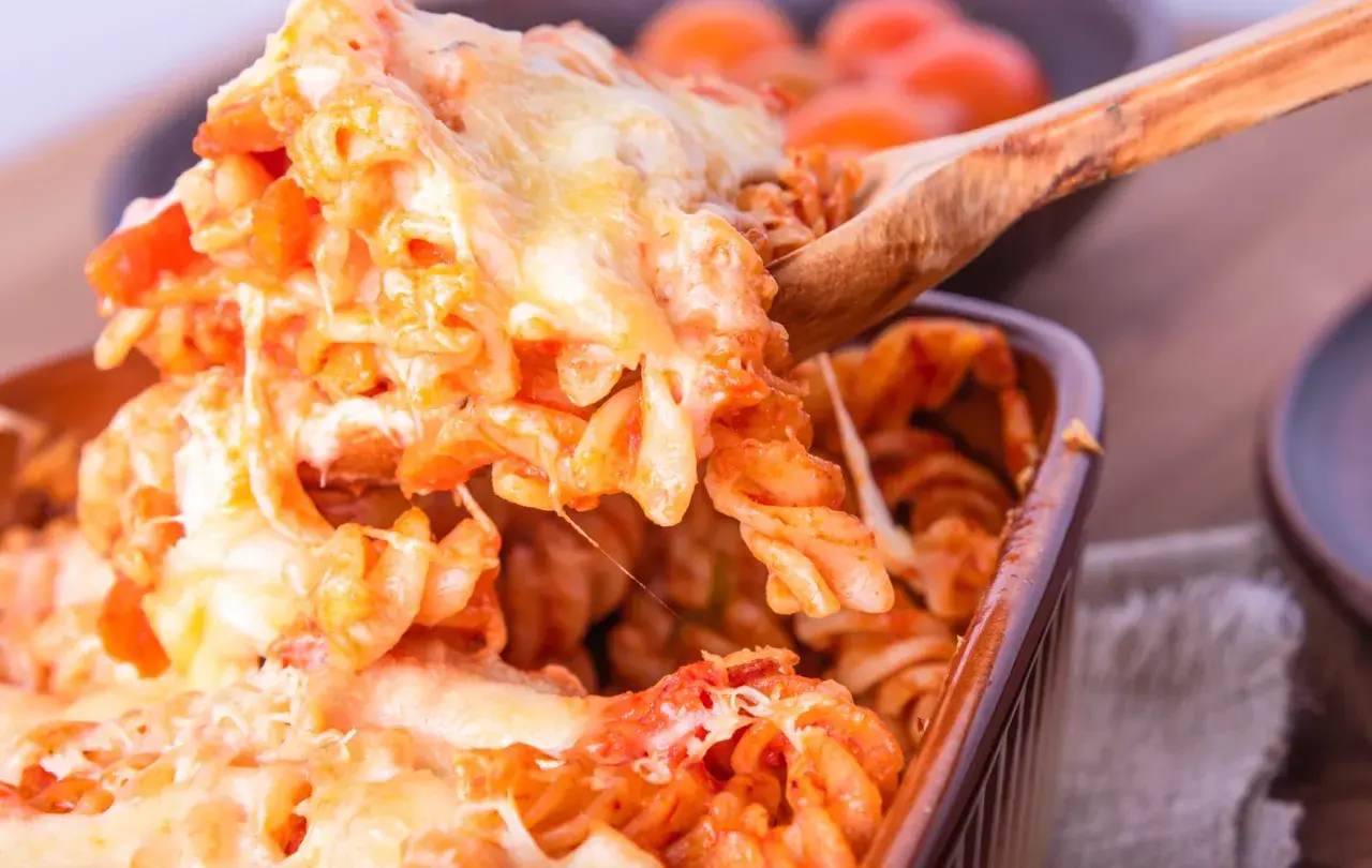 Un cucchiaio solleva una porzione generosa di pasta alla sorrentina, filante e dorata, da una pirofila marrone.