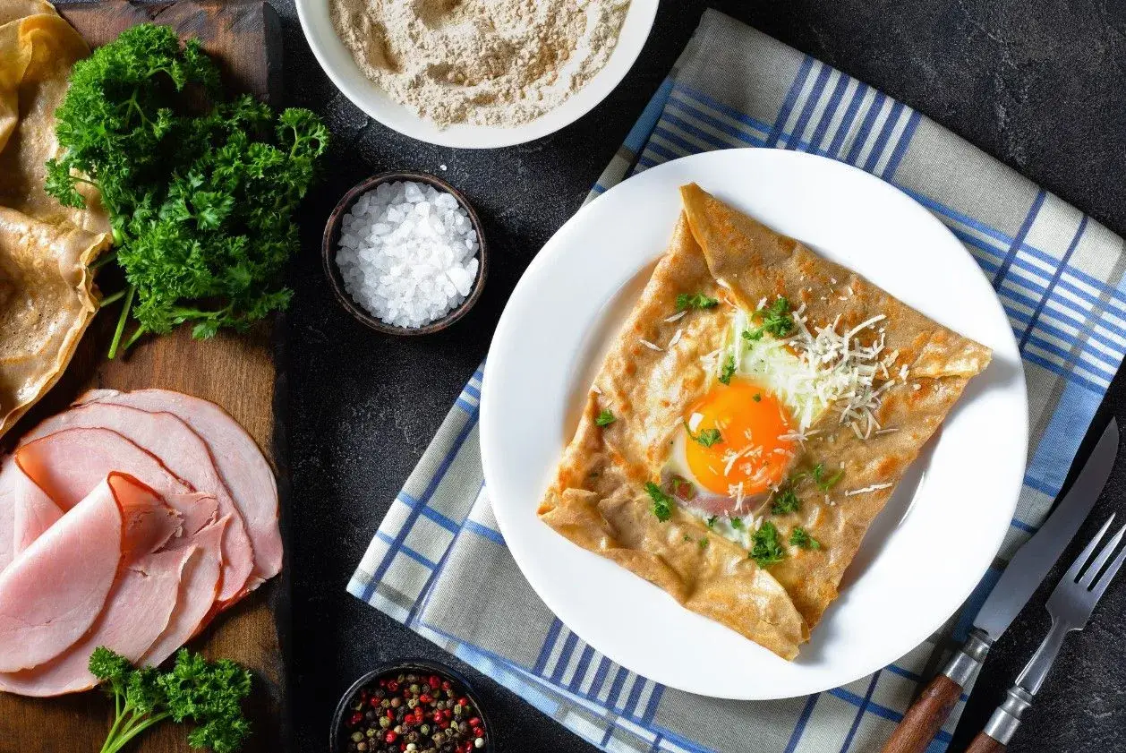 Francuska crepe z jajkiem sadzonym, serem i natką pietruszki na talerzu, obok szynka i składniki.