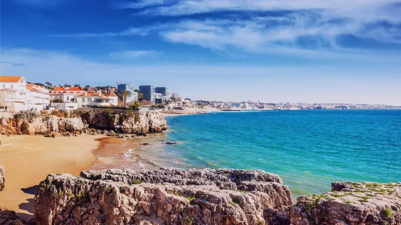 Felsige Küste mit Sandstrand und weißen Häusern, Blick auf das türkisfarbene Meer und die Skyline von Lissabon. Ein perfekter lissabon strand.