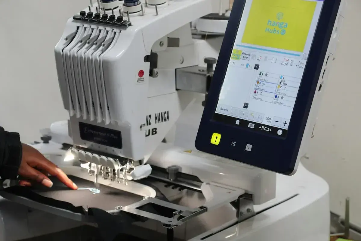 Une machine à broder professionnelle, type impression DTG, crée un logo sur un tissu. L'écran tactile affiche les détails du projet.
