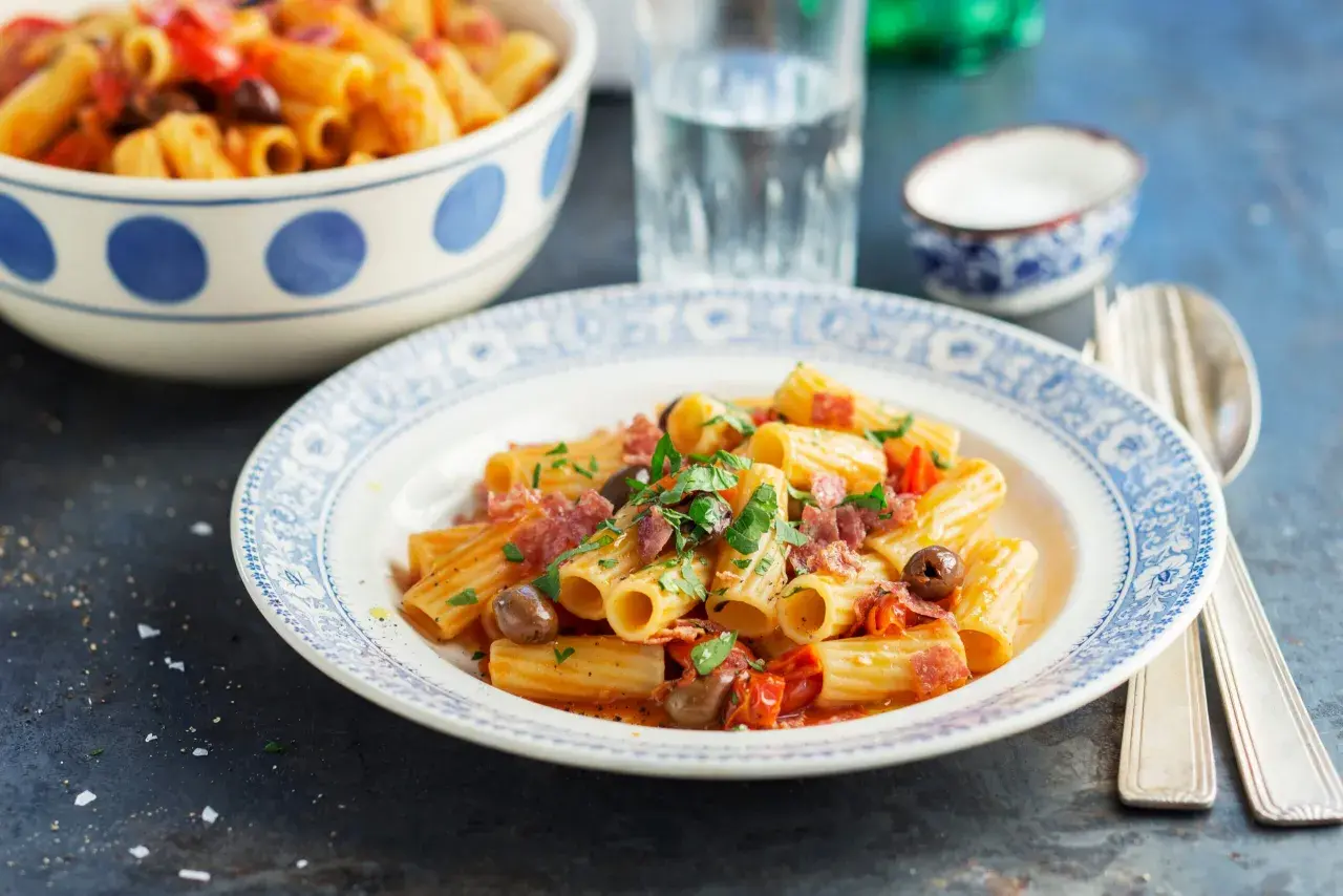 En tallrik med rigatoni pasta med salami, oliver och tomater, toppad med färsk persilja. En skål med mer pasta syns i bakgrunden.