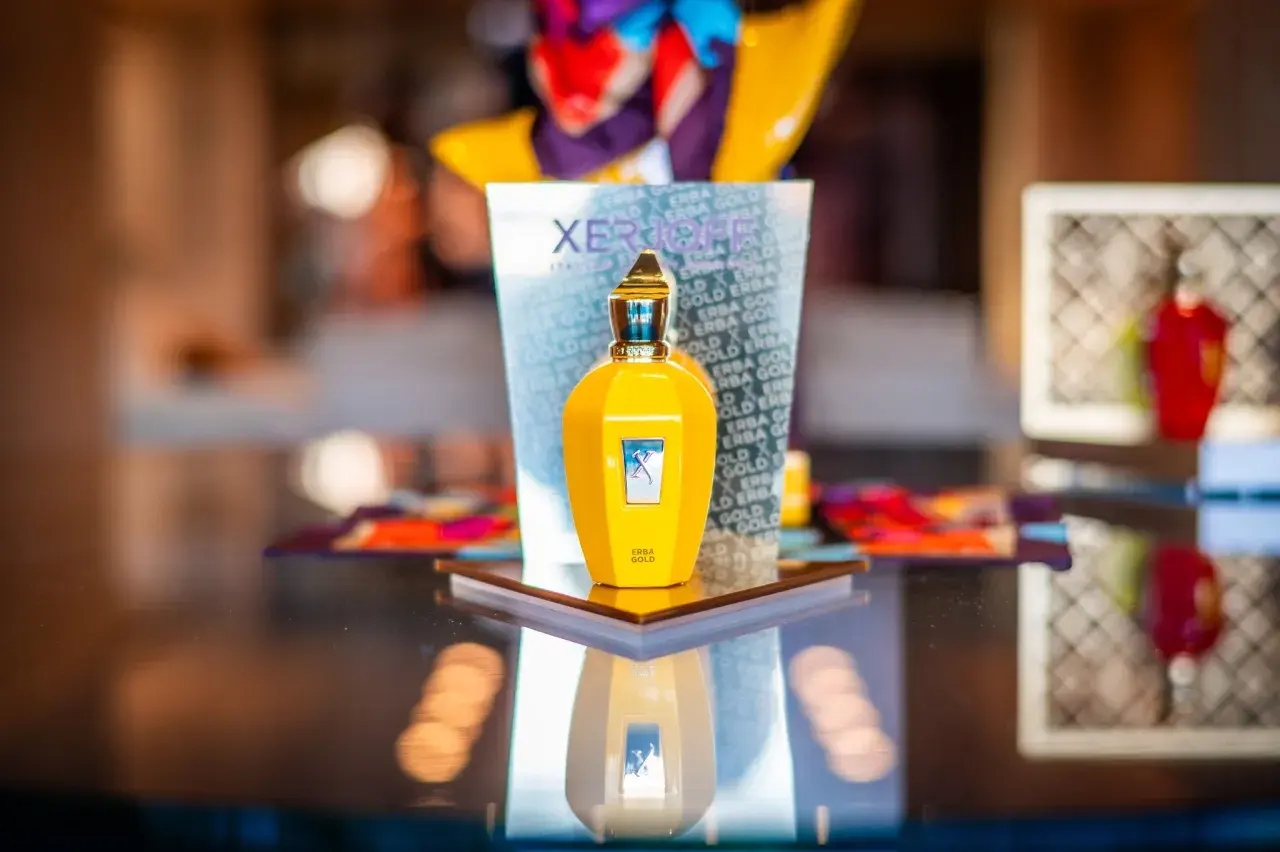 Żółta butelka perfum Xerjoff Erba Gold na lustrzanym blacie, odbijająca się w tafli.