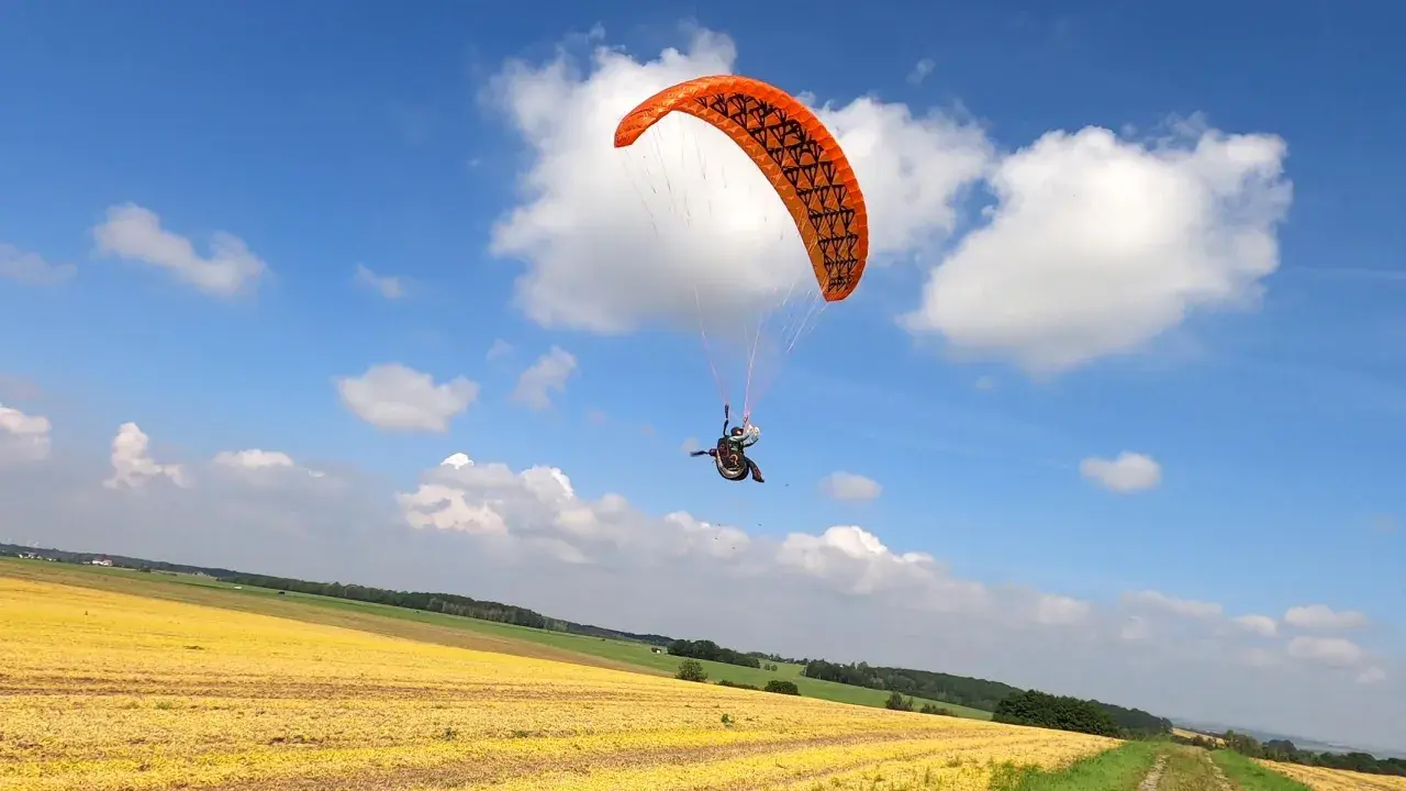 Ein Gleitschirmpilot mit einem orangen hepf micro vario Schirm fliegt über ein gelbes Feld unter blauem Himmel mit Wolken.