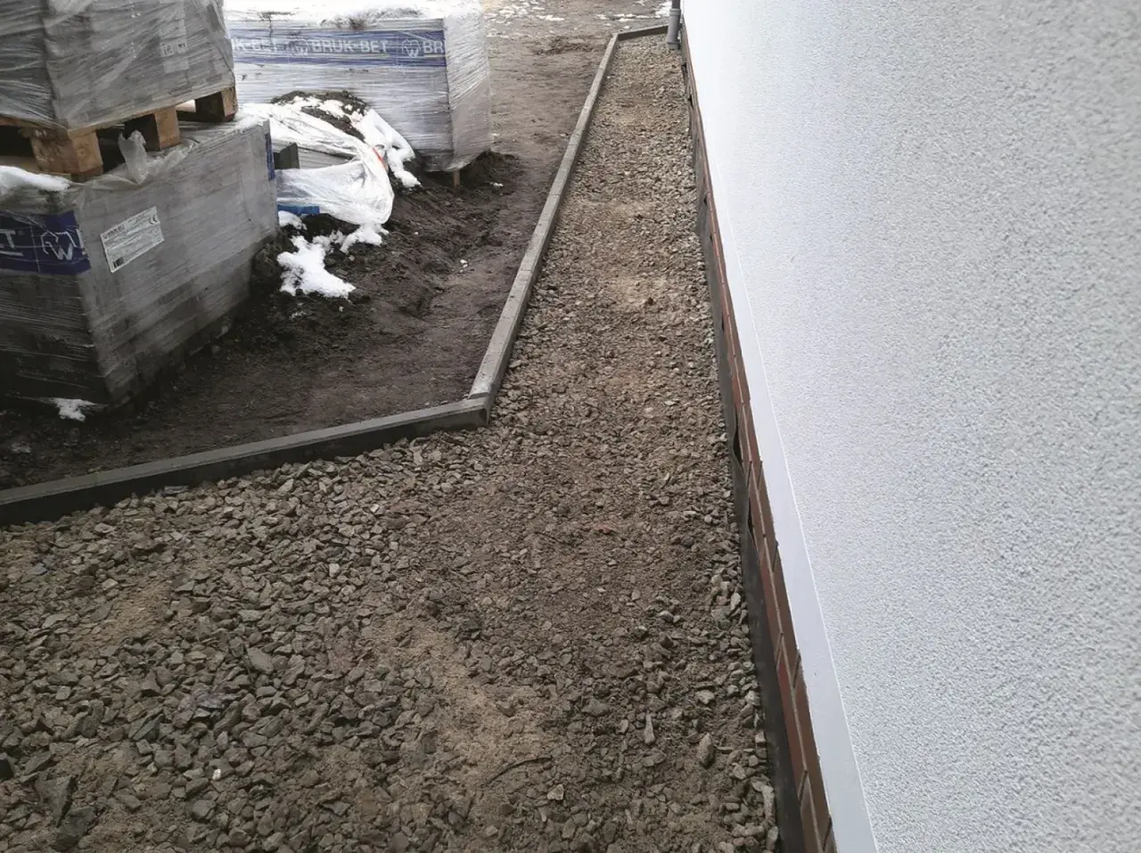 Niedokończony podjazd do domu, z paletami kostki brukowej i żwirem.
