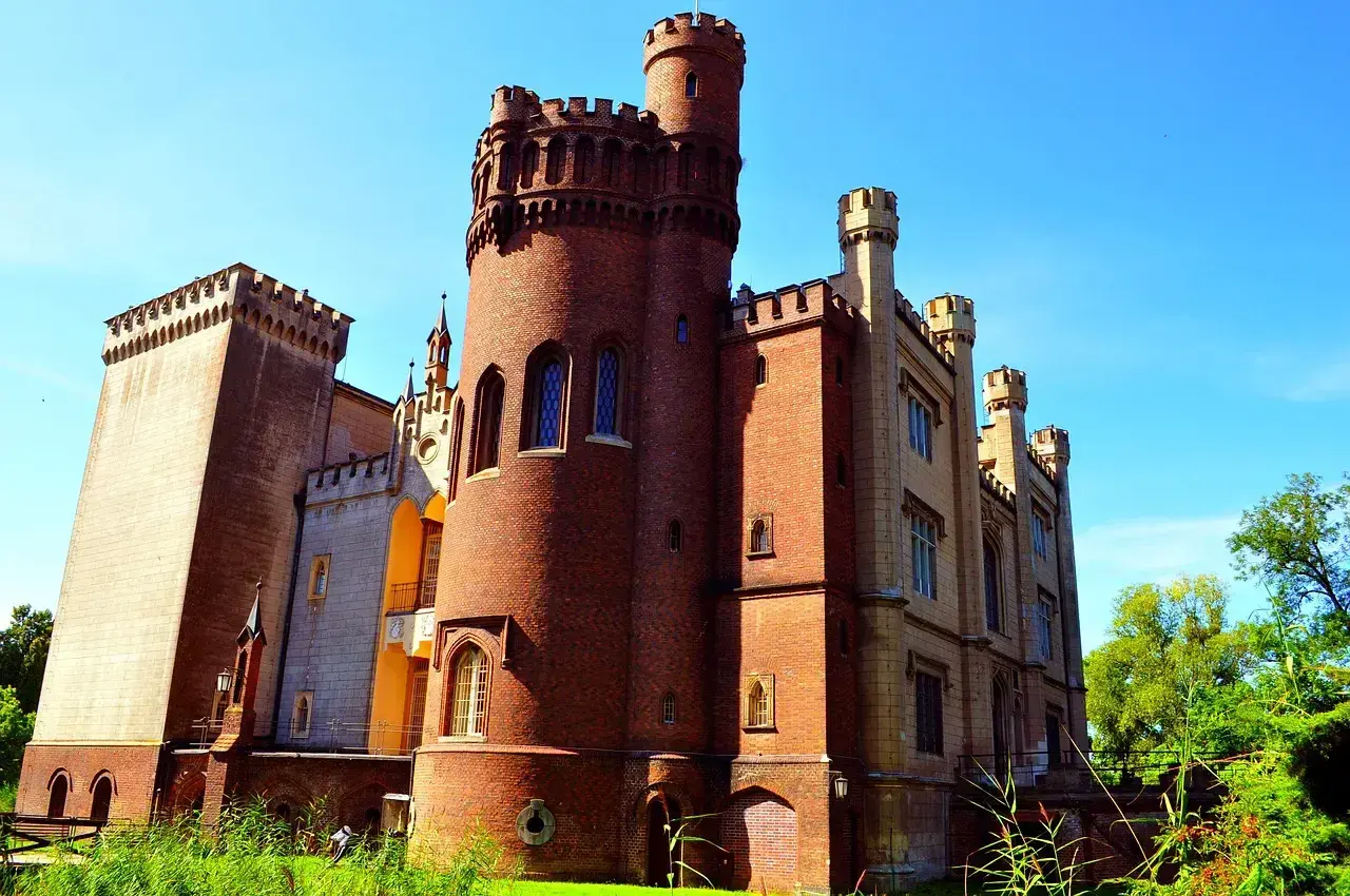 Zamek w Kórniku, z ceglaną wieżą i kamiennymi elementami, wznosi się dumnie na tle błękitnego nieba. Otoczony zielenią, zaprasza do odkrywania swoich tajemnic.