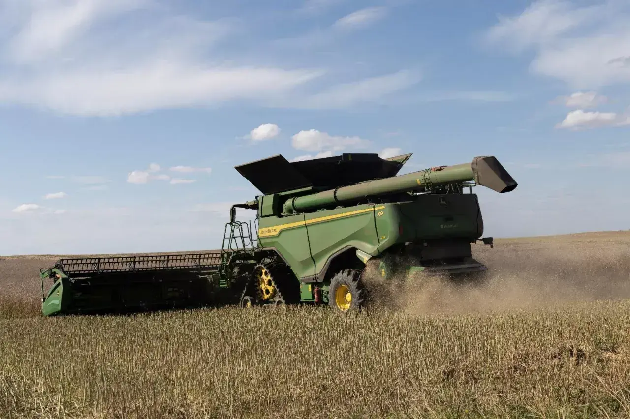 Zielona maszyna John Deere pracuje na polu, kiedy są żniwa. Widać kurz unoszący się z tyłu kombajnu.