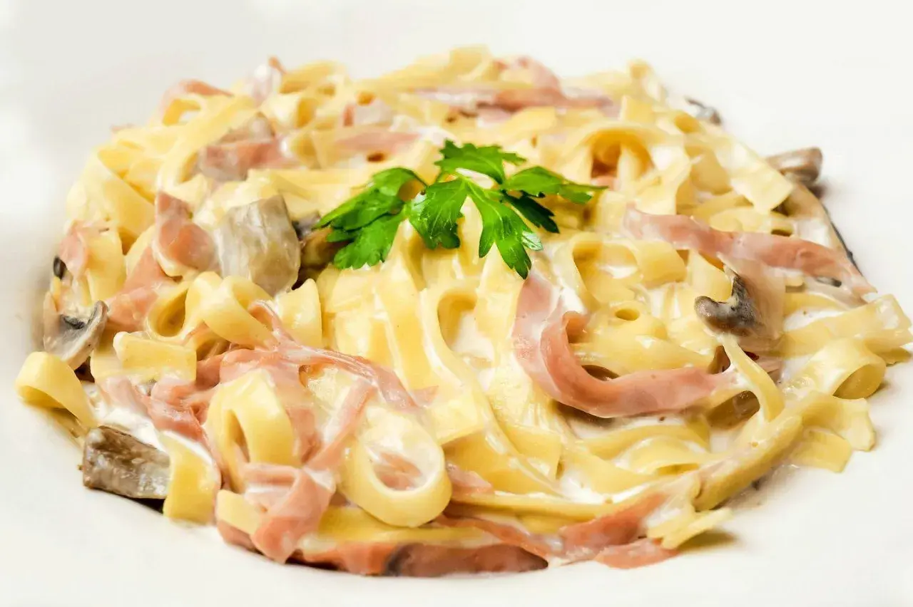 Tagliatelle z szynką i pieczarkami w kremowym sosie, z dodatkiem mozzarelli, udekorowane natką pietruszki.