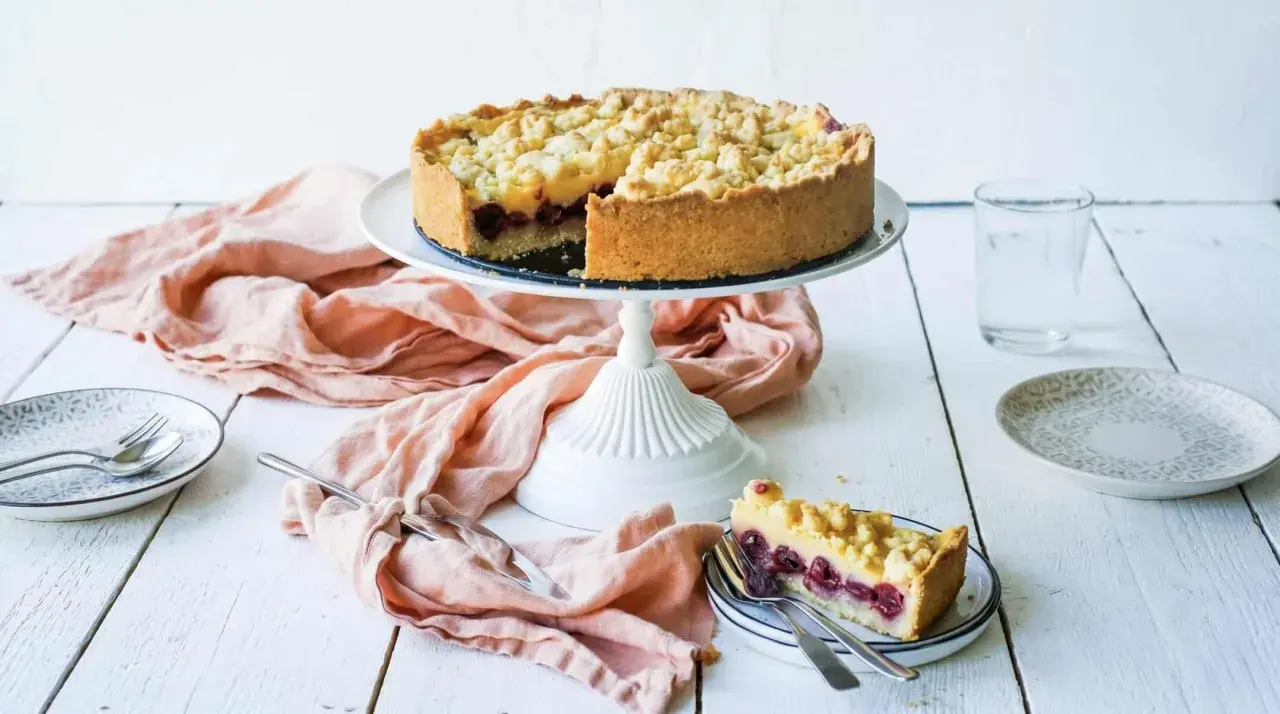 Ein saftiger Kirschkuchen mit Pudding und Streuseln auf einem Kuchenständer. Ein Stück ist bereits auf einem Teller angerichtet.