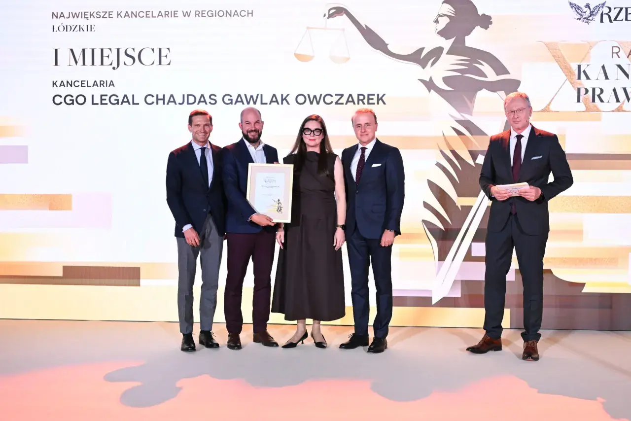 Pięć osób w garniturach i sukience stoi na scenie, odbierając nagrodę.