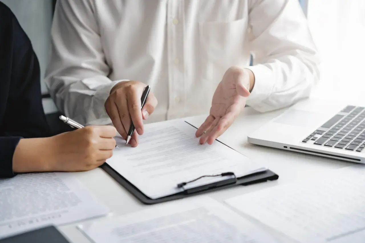 Deux personnes discutent d'un document, l'une signant, l'autre expliquant. Une commission d'intervention est en cours.