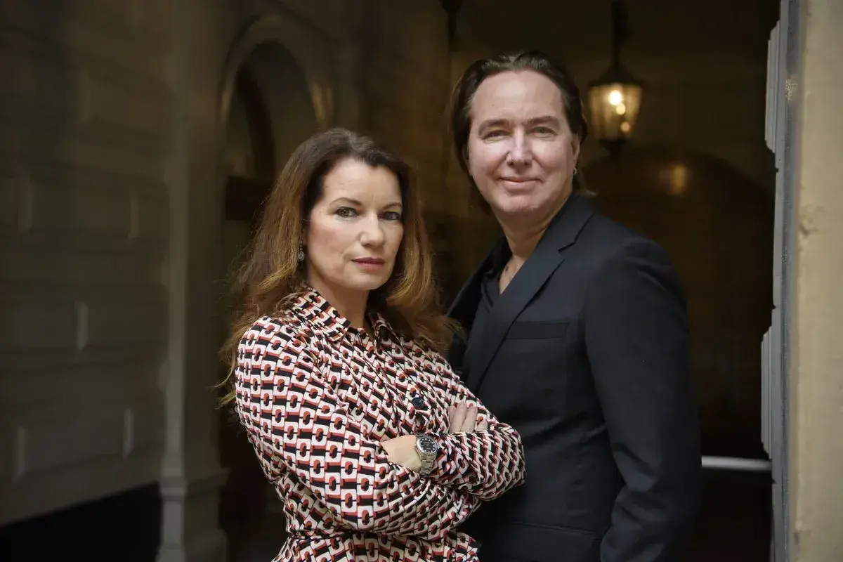 Alexander Ahndoril och en kvinna står nära varandra. Kvinnan bär en mönstrad blus och mannen en mörk kostym.