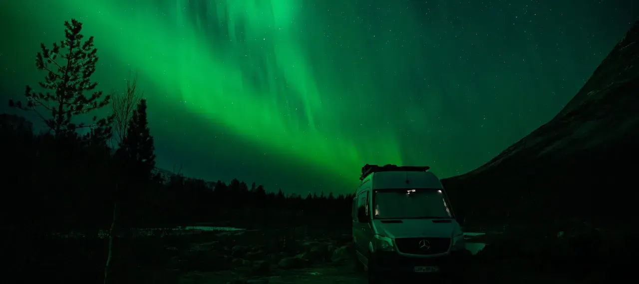 Ein Camper steht unter dem faszinierenden Licht der Aurora Borealis. Grüne Schleier tanzen am sternenklaren Himmel über dunklen Bäumen und einer Bergflanke.