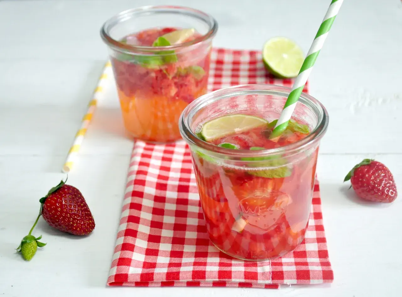 Orzeźwiający drink z truskawkami, miętą i limonką, podany w szklance z zielono-białą słomką. Obok leży świeża truskawka.