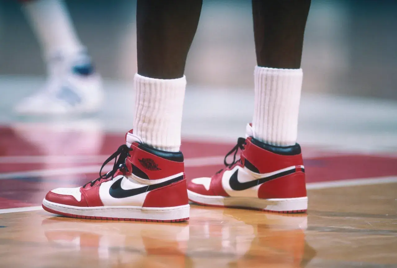 Rote und weiße Air Jordan 1 Sneaker mit schwarzem Nike-Logo auf einem Basketballplatz. Ein ikonisches Stück der Geschichte des Nike-Unternehmens.