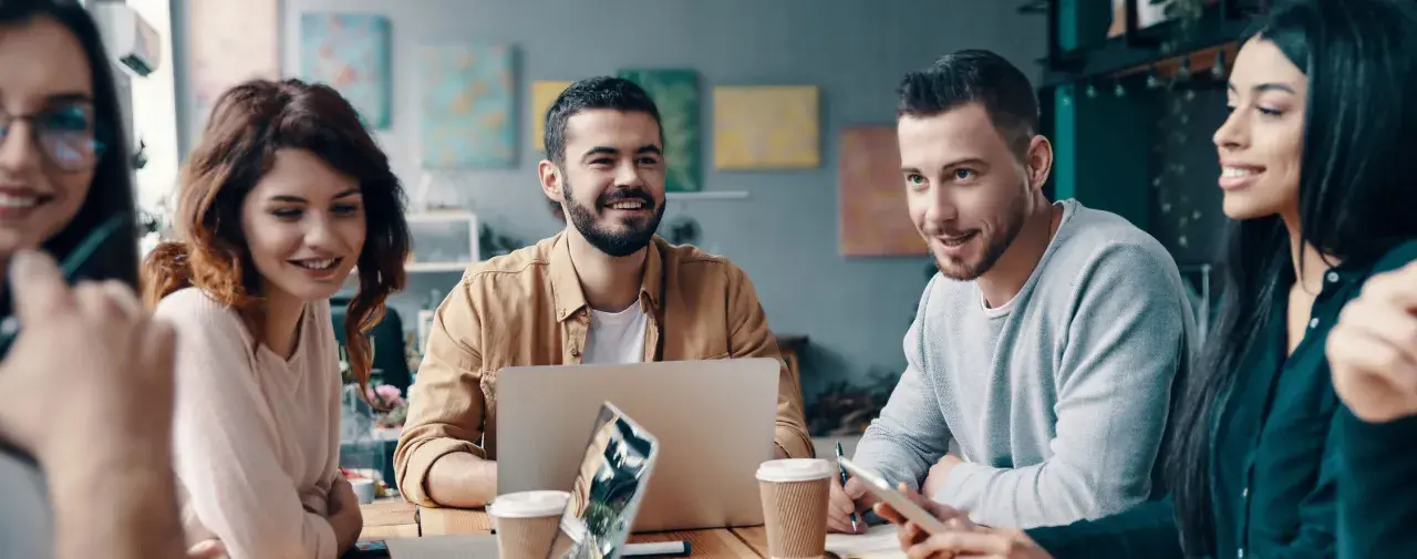En grupp unga, leende människor diskuterar vid ett bord. En laptop och kaffekoppar antyder ett möte om utbildning, organisation och ledarskap.