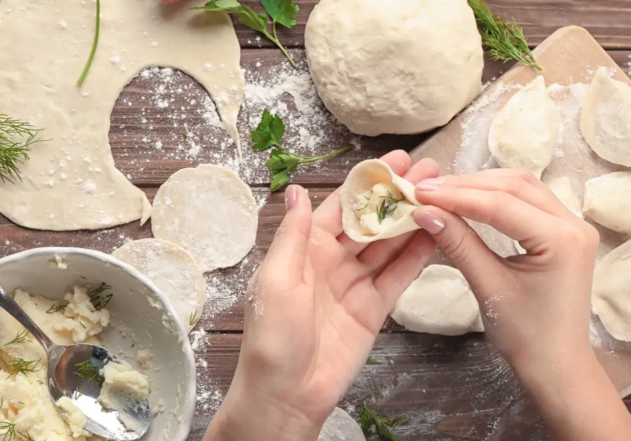 Ręce formują pieroga z ciasta na pierogi, wypełnionego farszem ziemniaczanym i koperkiem. Na drewnianym blacie widać mąkę, gotowe pierogi i miskę z farszem.