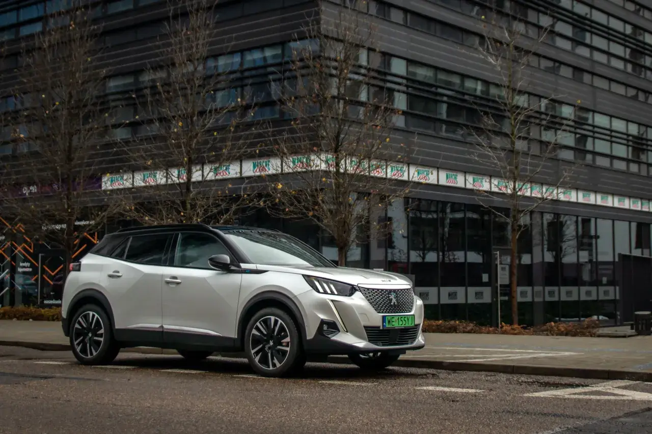 Biały Peugeot e-2008 zaparkowany przed nowoczesnym budynkiem.