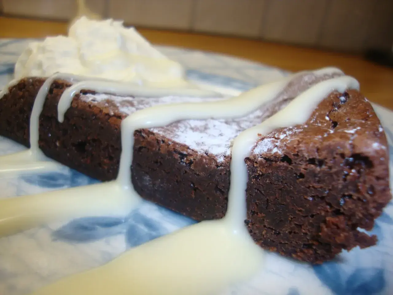 En bit mörk kladdkaka med pudersocker och vit chokladsås, serverad med vispad grädde.