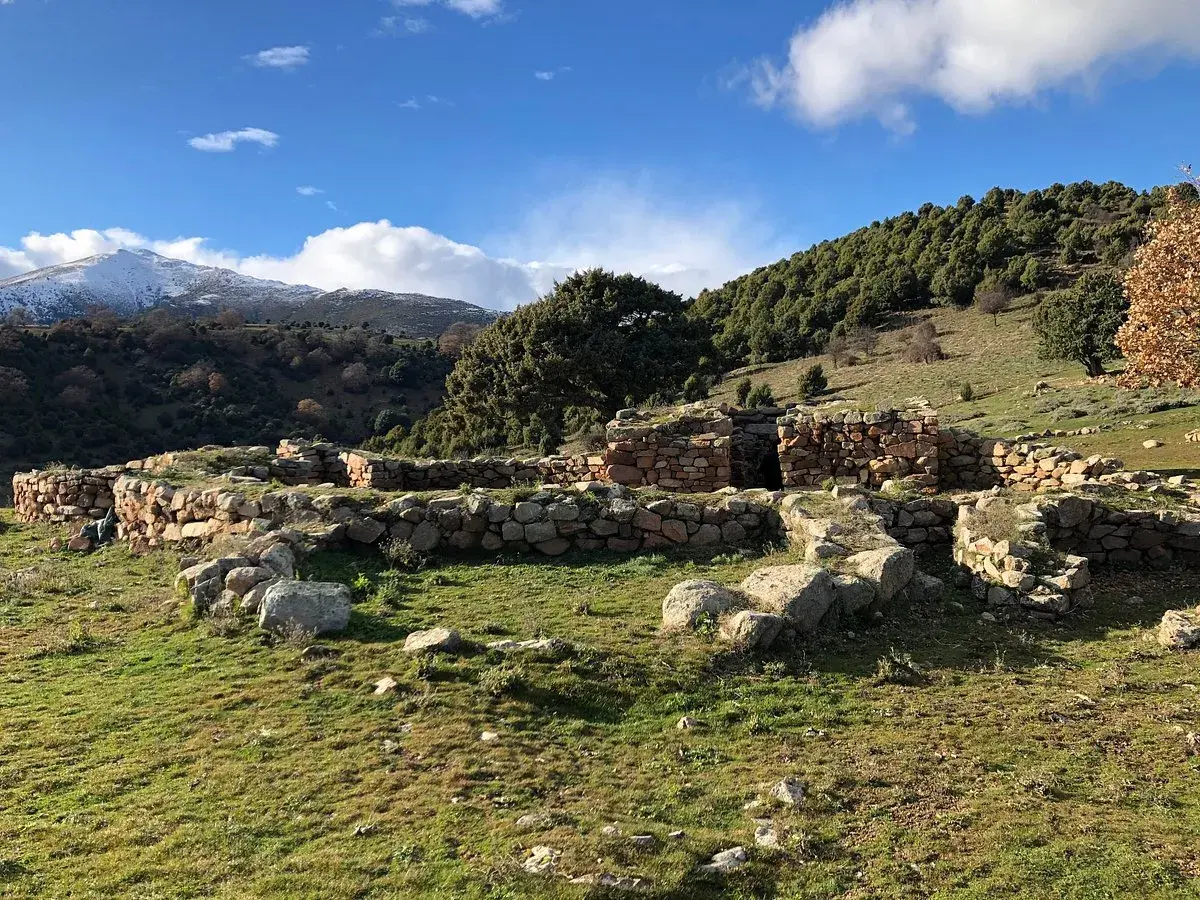 Antiche rovine di pietra, s'arcu e is forros, su un prato verde sotto un cielo azzurro con nuvole bianche e montagne innevate sullo sfondo.