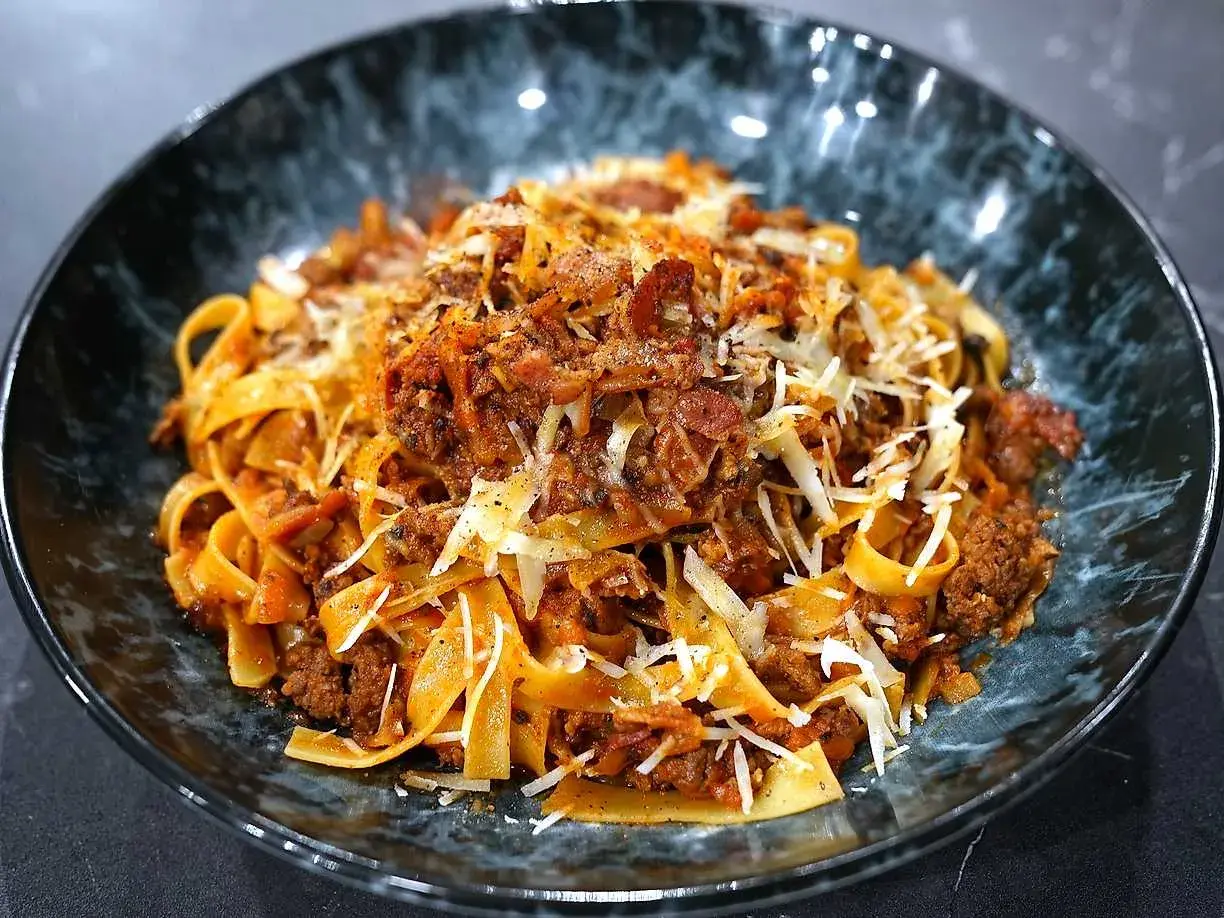 En skål med breda bandnudlar täckta av en mustig köttfärssås, toppad med riven ost och peppar. En härlig pasta ragu!