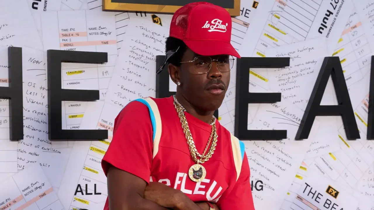 Tyler, The Creator w czerwonej czapce i naszyjnikach, na tle liter "HE" i notatek.
