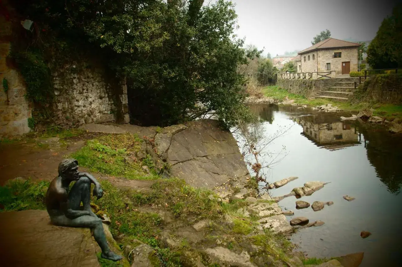 Escultura de hombre pensativo junto a un río, con una casa de piedra reflejada en el agua. Un lugar tranquilo que ver cerca de Liérganes.