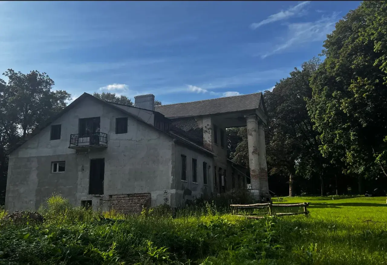 Zniszczony dworek z kolumnami i balkonem, otoczony zielenią.