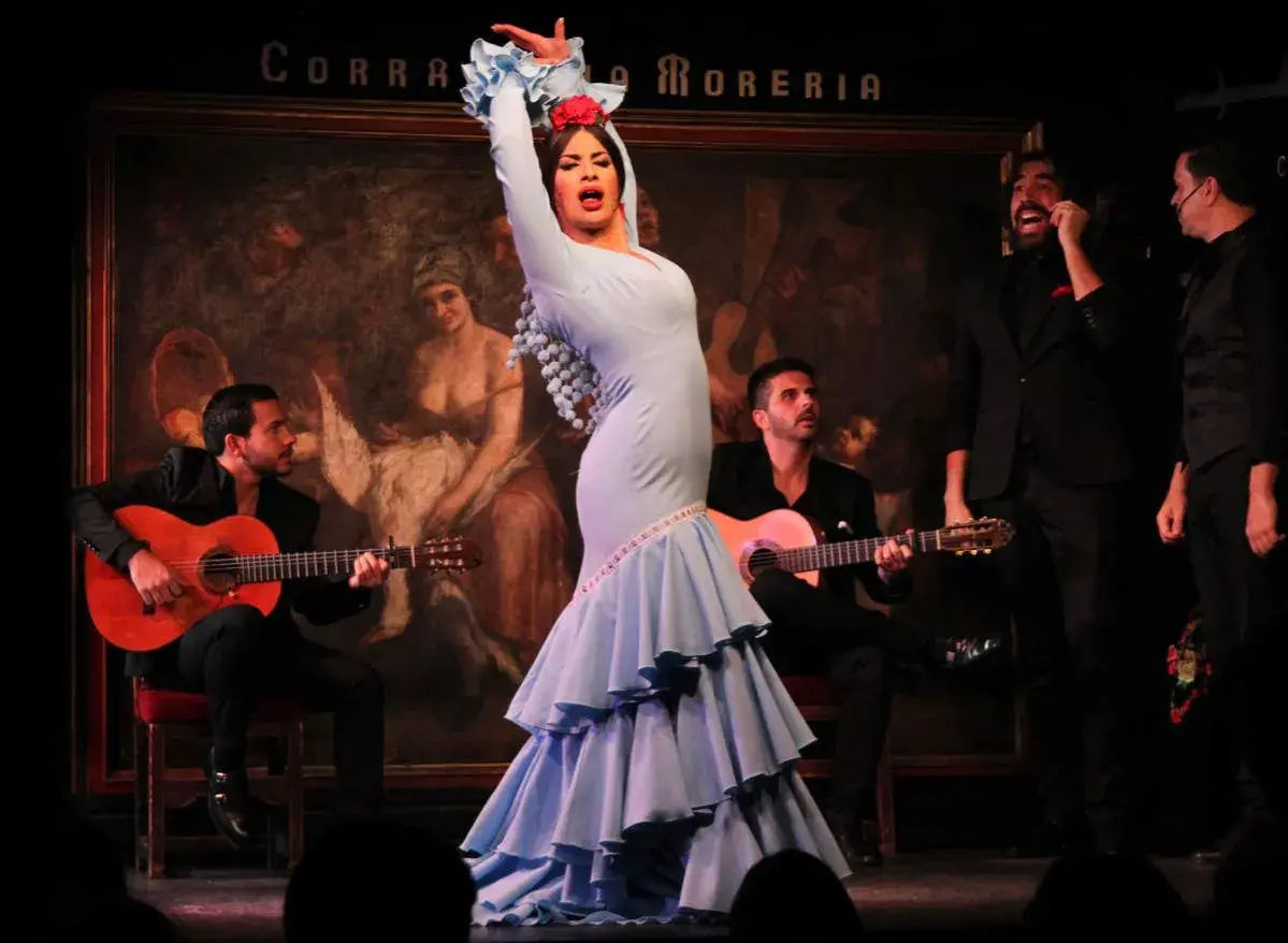 La mejor bailaora de flamenco, con un vestido azul voluminoso, cautiva en el escenario mientras dos guitarristas la acompañan.
