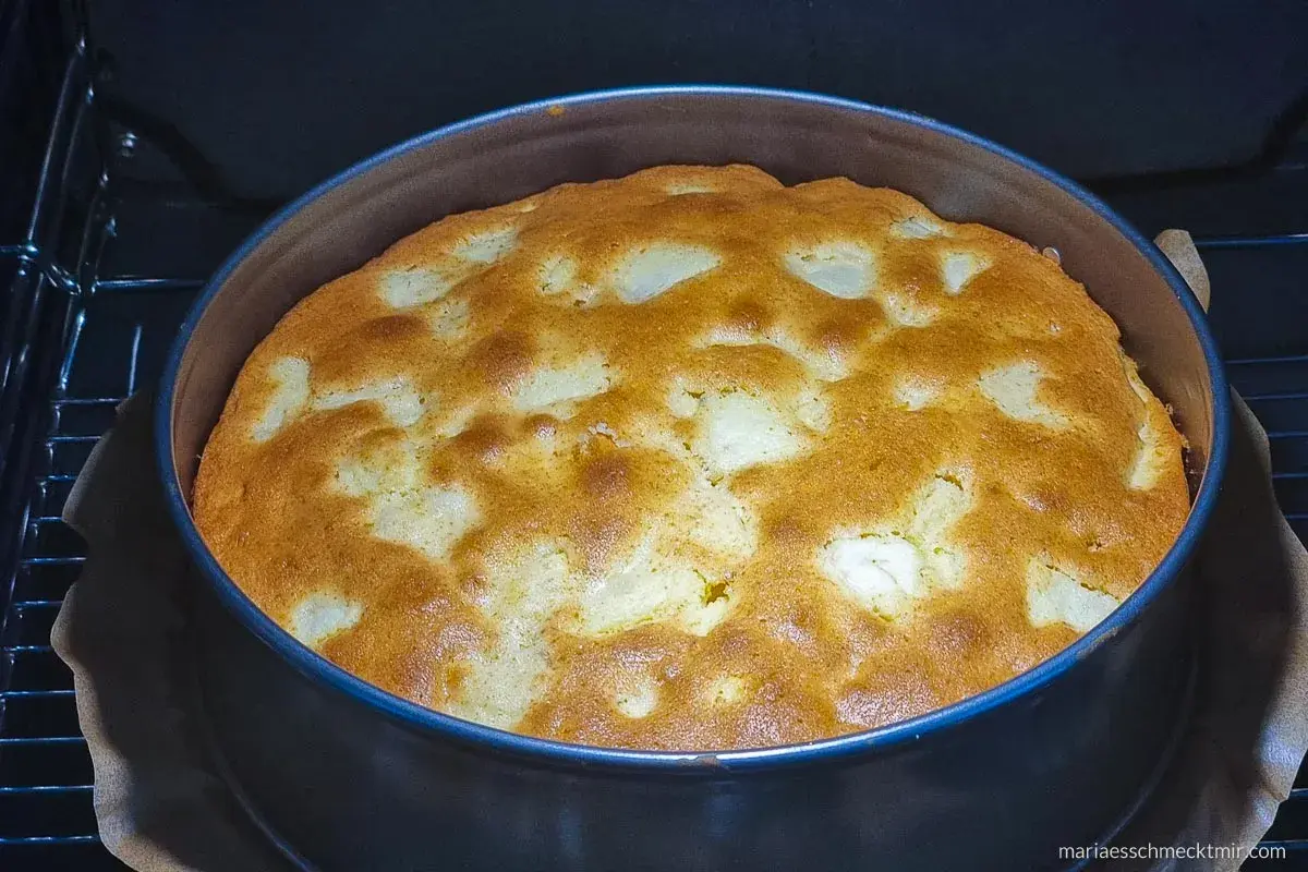 Ein goldbrauner, versunkener Apfelkuchen bäckt in einer runden Form im Ofen. Die Äpfel sind im Teig versunken und bilden ein schönes Muster.