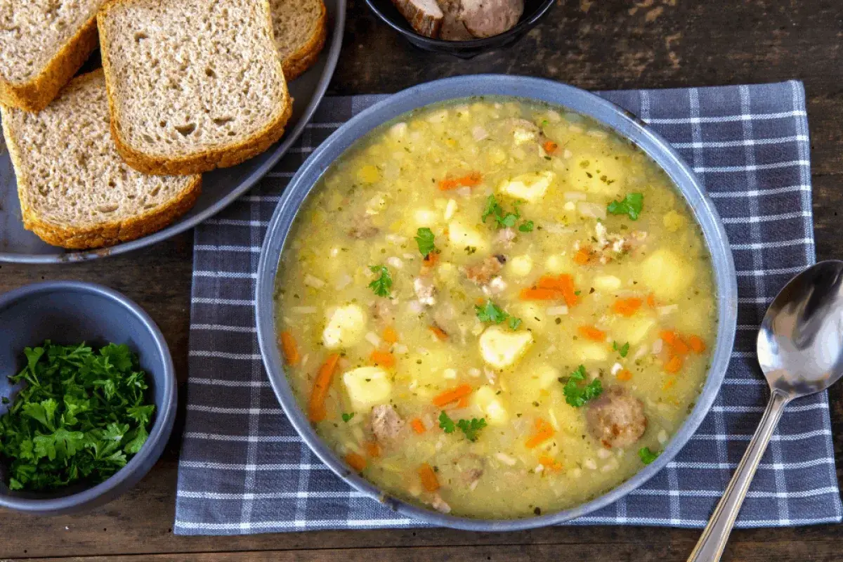 Gęsta zupa grochowa z ziemniakami, marchewką i kiełbasą. Obok kromki chleba i miseczka natki pietruszki – idealne dodatki do zupy grochowej.