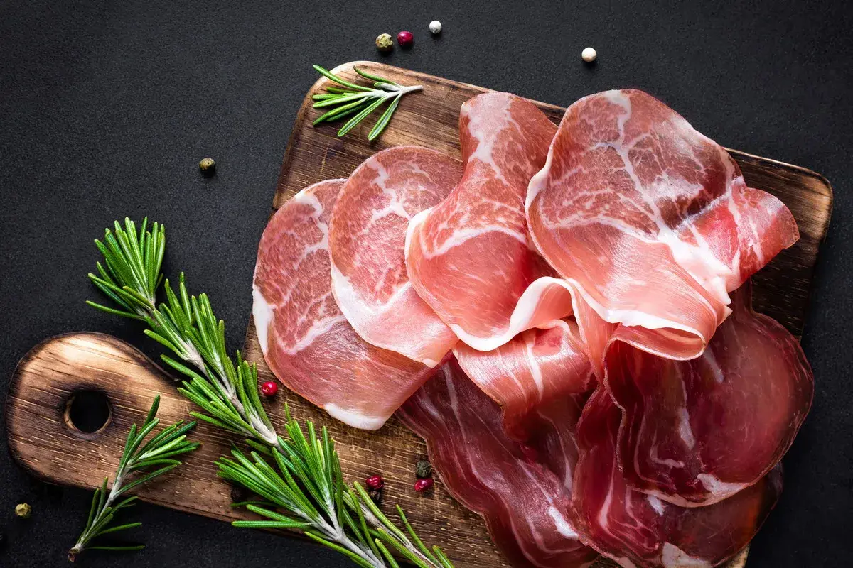Cienkie plastry prosciutto, w tym szynka parmeńska, ułożone na drewnianej desce z gałązkami rozmarynu i ziarnami pieprzu.
