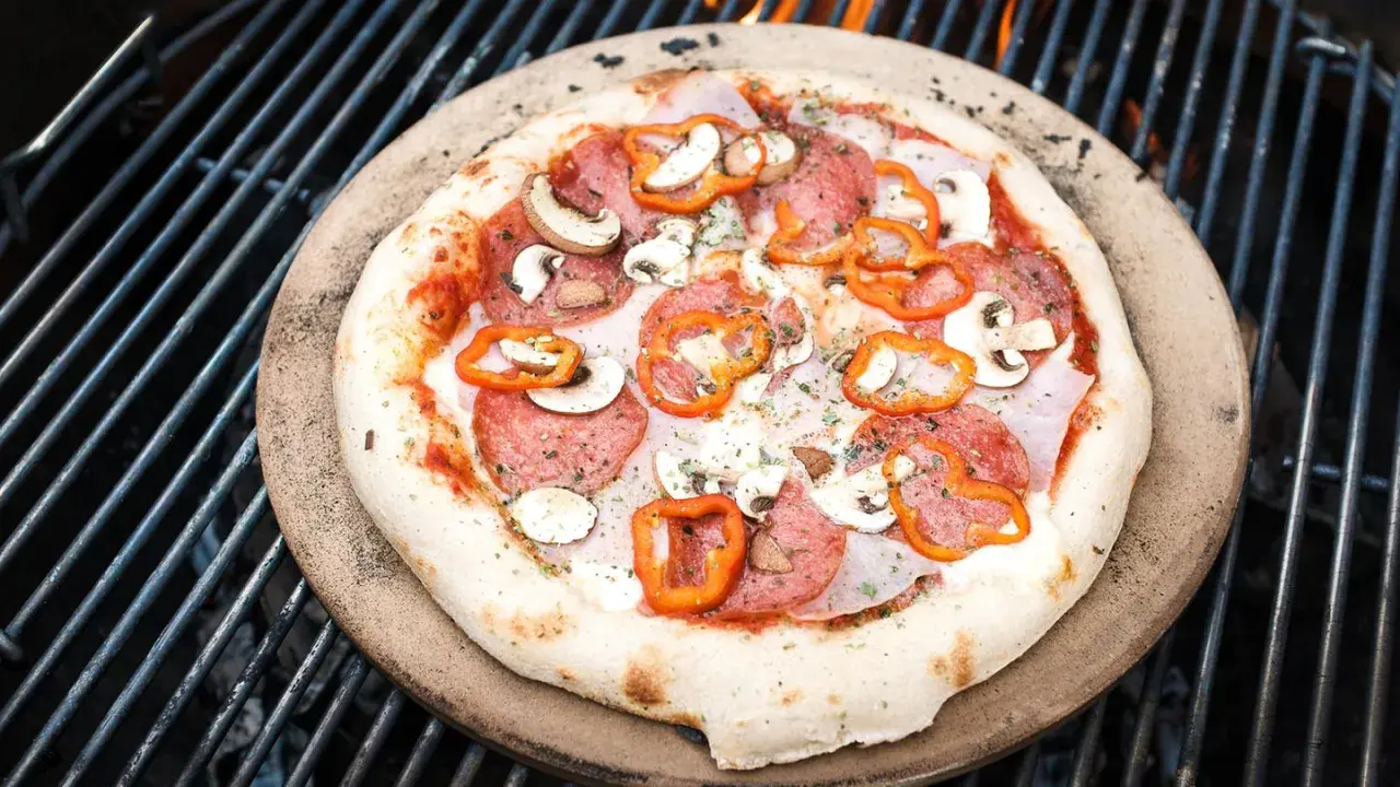 Eine köstliche Pizza mit Salami, Pilzen und Paprika wird auf einem Pizzastein über glühenden Kohlen auf dem Grill zubereitet.