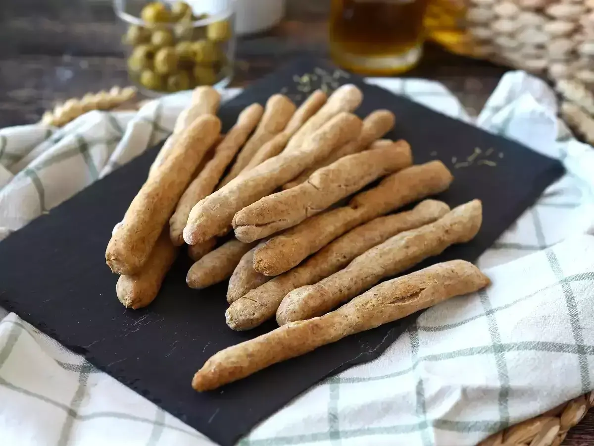 Une pile de gressins dorés, parfaits pour une recette gressin maison. Des olives et une boisson en arrière-plan.