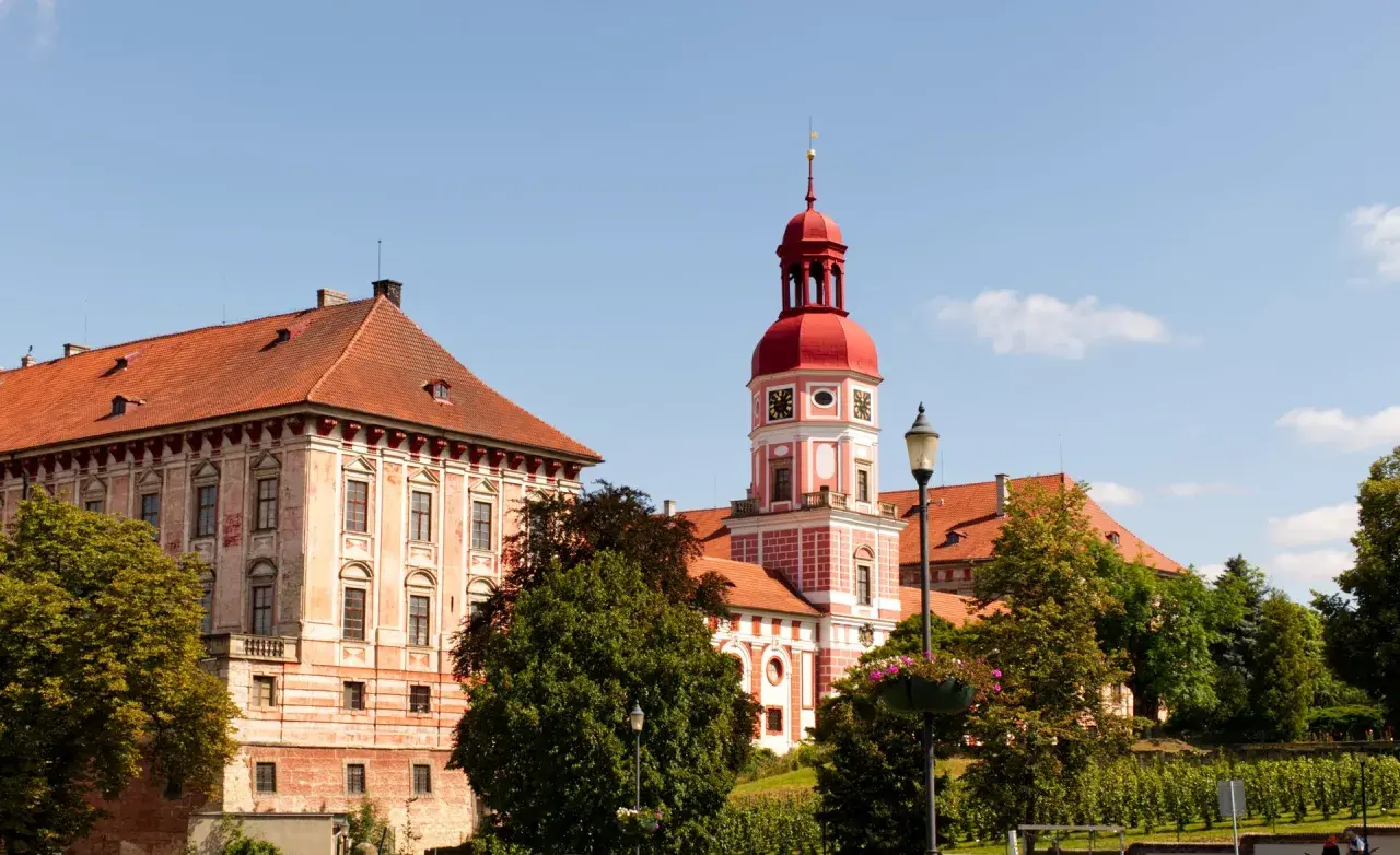 Schloss Roudnice nad Labem mit seinem roten Turm und den Ziegeldächern, umgeben von grünen Bäumen.
