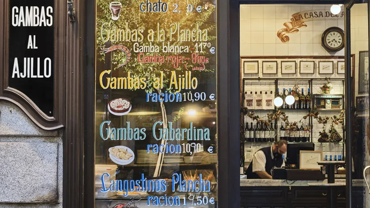 Cartel de un restaurante con menú de gambas y langostinos, y un camarero detrás del mostrador.