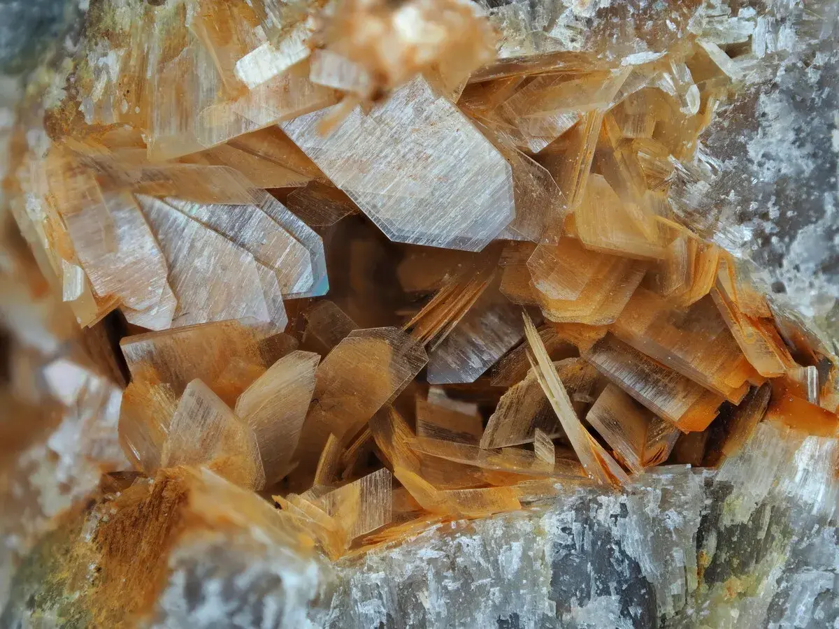 Nahaufnahme von Anhydrit-Kristallen, die wie dünne, schichtartige Plättchen in warmen Orangetönen und transparenten Farben angeordnet sind.