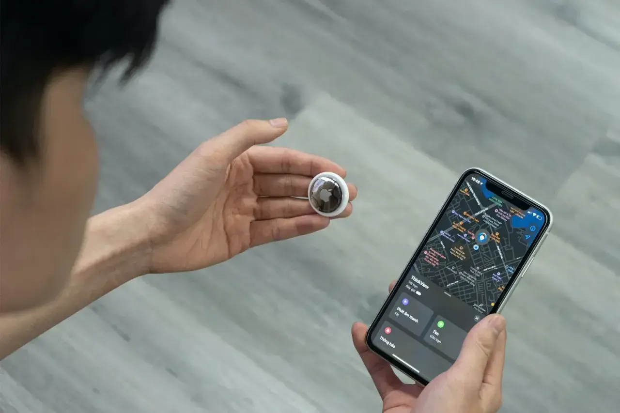 Osoba trzymająca w dłoni lokalizator AirTag i iPhone'a z mapą. AirTag co to? To małe urządzenie do śledzenia zgubionych przedmiotów.