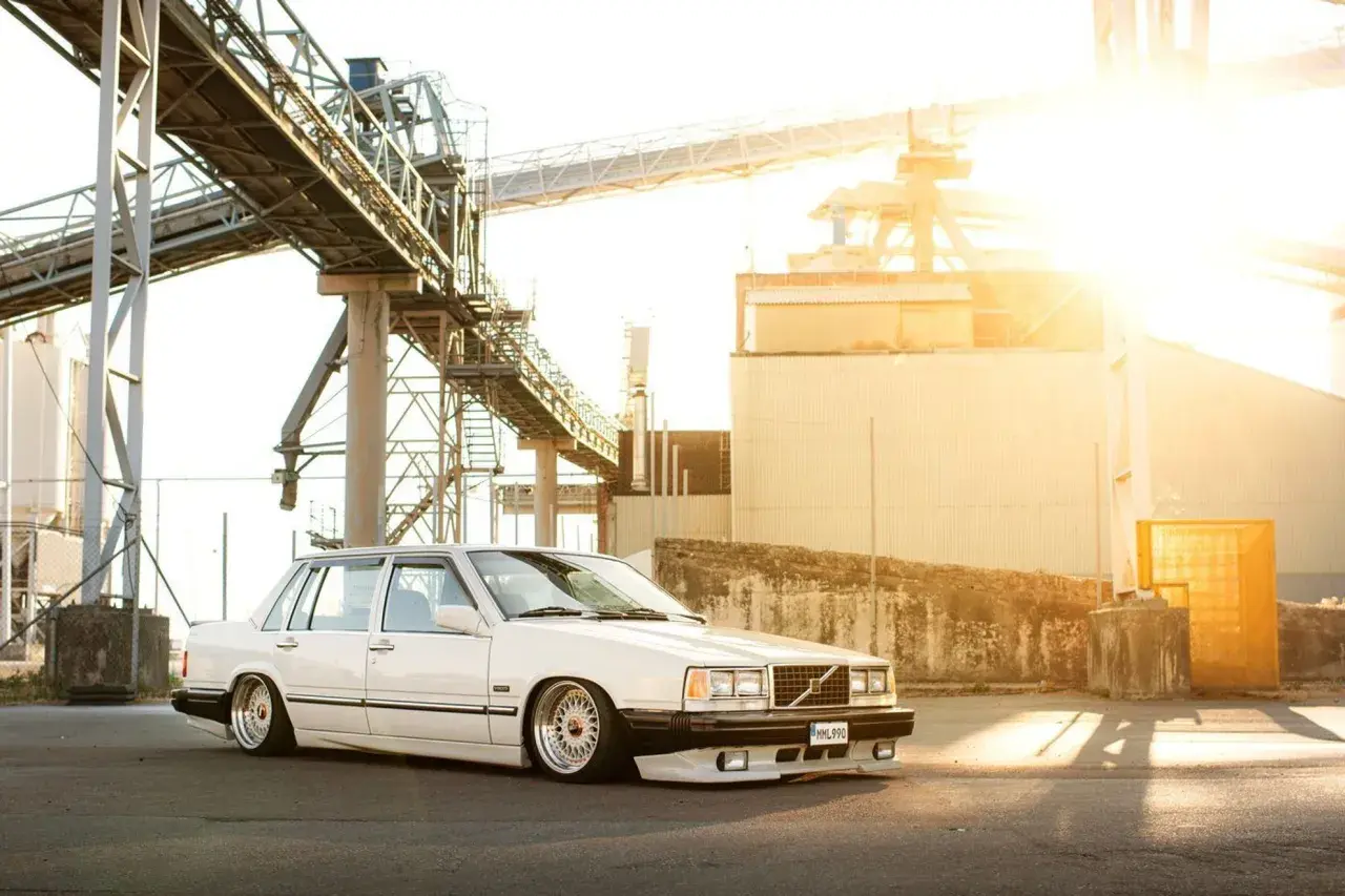 Vit Volvo 740 från 1990, sänkt och med kromade fälgar, parkerad vid en industrianläggning i solnedgången.
