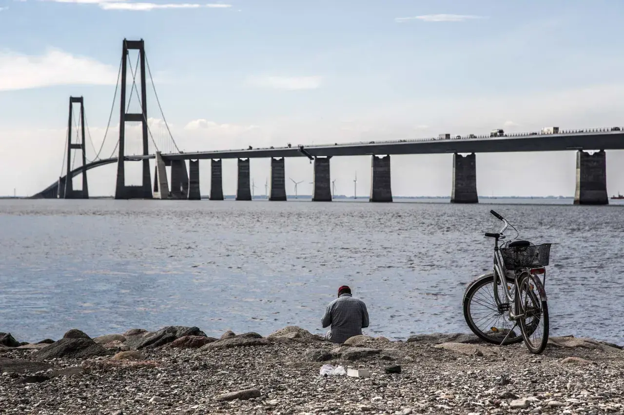En man sitter vid vattnet med sin cykel, blickandes mot den imponerande Stora Bältbron.