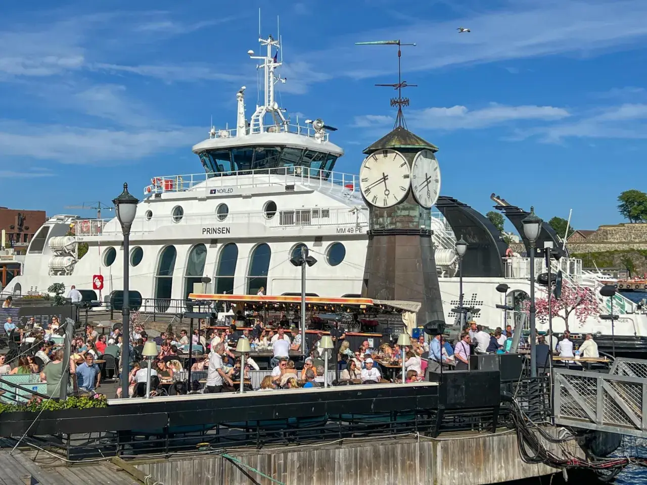 Tętniący życiem deptak w Oslo, z ludźmi siedzącymi w kawiarniach, na tle białego statku i zabytkowej wieży zegarowej. Oslo czy warto? Zdecydowanie tak!