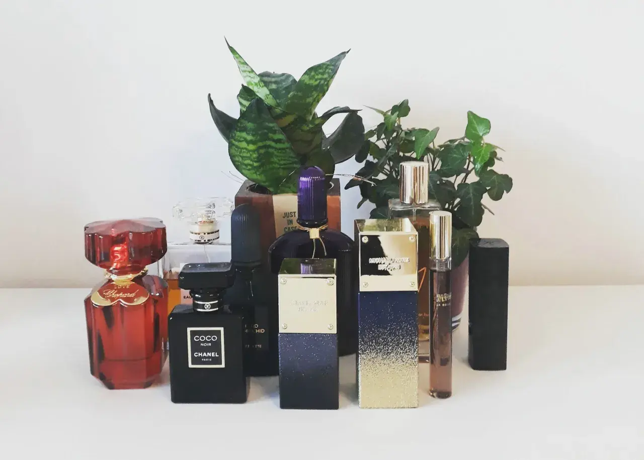Kolekcja perfum: czerwony Chopard, czarny Coco Noir Chanel 100ml, fioletowy i złoty Michael Kors, oraz inne, otoczone zielenią.