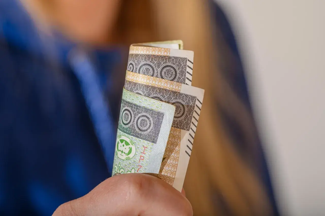 Orzeczenie o niepełnosprawności to podstawa do ubiegania się o zasiłek pielęgnacyjny. Dłonie trzymają banknoty 100 zł.