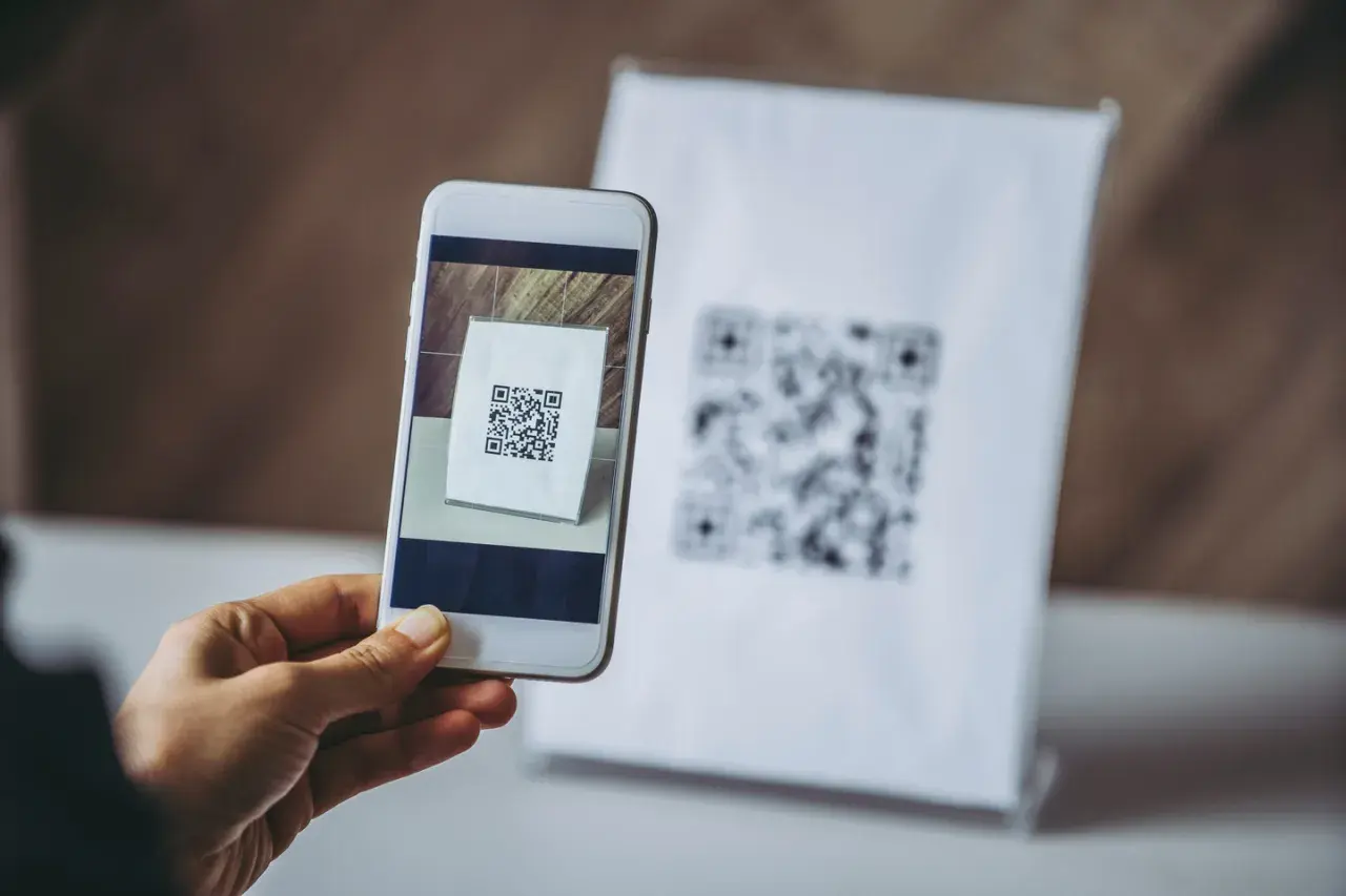 Dłoń trzyma smartfon skanujący kod QR na białej kartce.