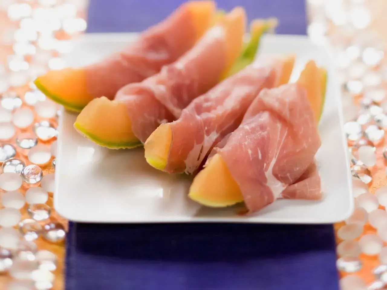 Saftige Honigmelone mit zartem Schinken umwickelt, serviert auf einem weißen Teller. Ein süß-salziger Genuss!