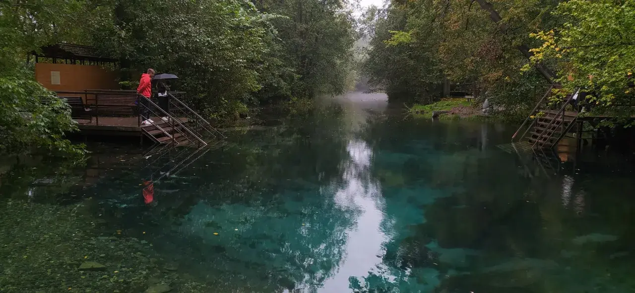 Turkusowe jezioro w Rosji, otoczone drzewami. Dwie osoby stoją na schodach prowadzących do wody, jedna z parasolem.