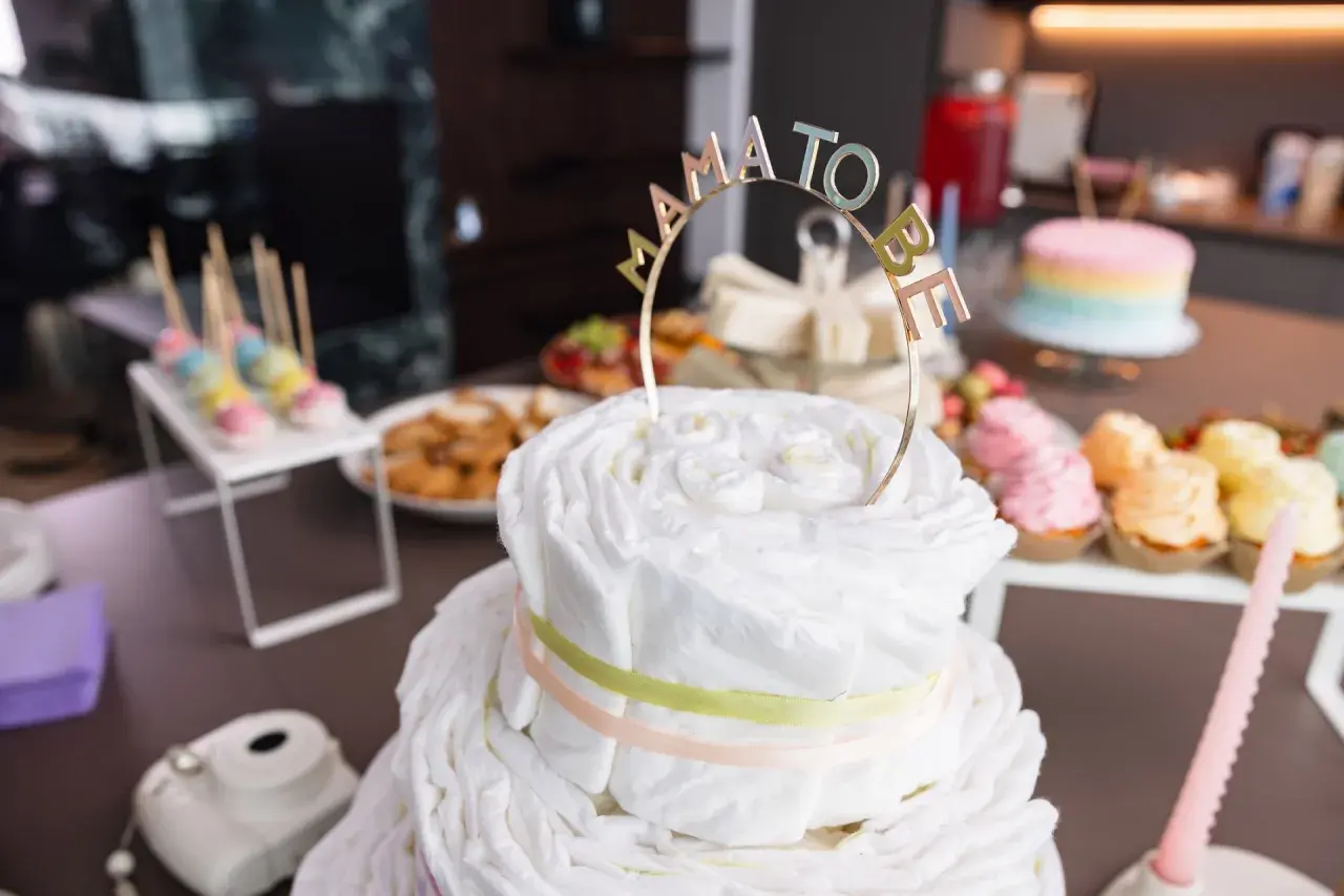 Una torta di pannolini decorata con un topper "Mama to Be" per un baby shower. Sul tavolo anche cupcakes colorati e dolcetti.