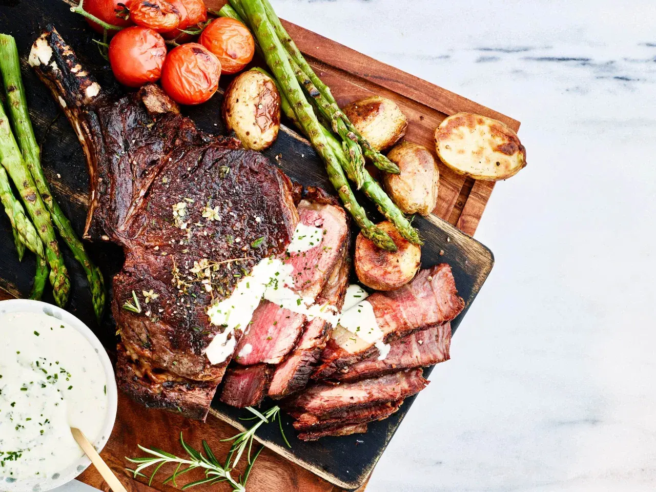 En saftig tomahawk-biff, perfekt grillad, serveras med rostade tomater, sparris och potatis. En krämig sås fulländar denna grillupplevelse.