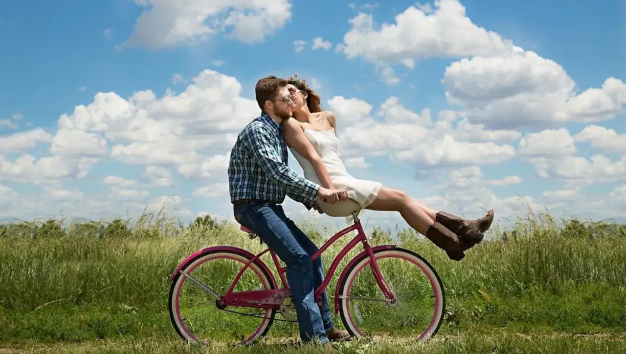 Coppia su bici rosa, lei seduta davanti a lui, baci appassionati sotto un cielo azzurro. Ecco perchè ci si innamora.