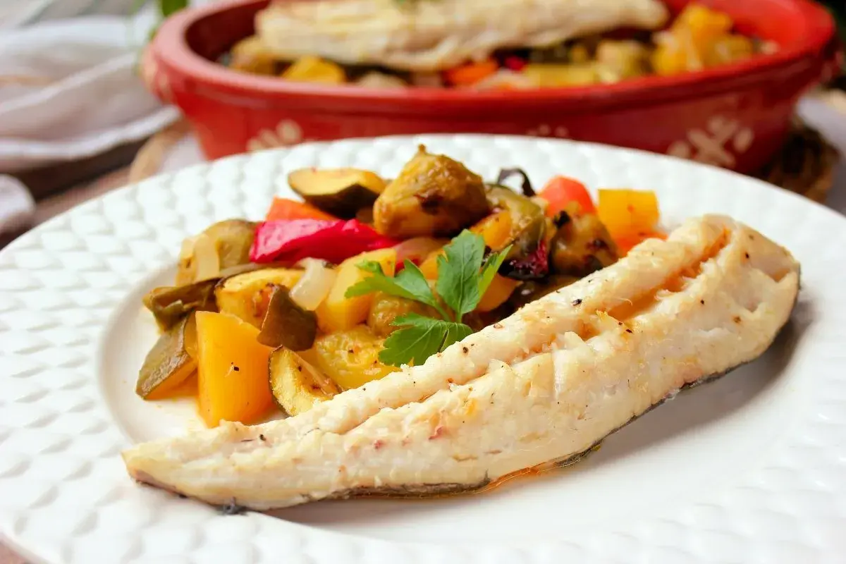 Filete de merluza al horno con verduras asadas, como pimientos, calabacín y patatas. Un plato saludable y delicioso.